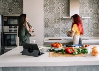 Contenuto giovani ragazze omosessuali con bottiglia di vino parlando mentre si guardano tra stufa e verdure fresche — Foto stock