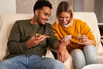 Содержание многорасовой пары, сидящей в гостиной во время еды пиццы и просмотра смешного видео на смартфоне в выходные дни — стоковое фото