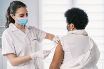 Especialista médica feminina em uniforme de proteção, luvas de látex e máscara facial vacinando a paciente madura afro-americana na clínica durante o surto de coronavírus — Fotografia de Stock