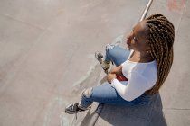 De cima fria fêmea preta com penteado trançado e em patins sentados na rampa no parque de skate e olhando para longe — Fotografia de Stock