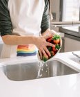 Cultiver jeune femelle dans le tablier laver les fraises fraîches sous l'eau liquide du robinet dans la cuisine maison — Photo de stock