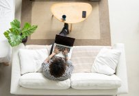 Jovem hipster macho com cabelos cacheados navegando na internet no netbook enquanto descansa no sofá na sala da casa — Fotografia de Stock