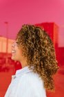 Vista lateral de la mujer afroamericana encantada con peinado rizado de pie sobre fondo rojo en el estudio mirando hacia otro lado - foto de stock