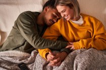 Высокий угол любящей многонациональной пары, расслабляющейся на диване под одеялом, обнимаясь и держась за руки — стоковое фото