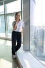 Jovem empresária de pé no escritório com grandes janelas tendo um telefonema no telefone celular — Fotografia de Stock