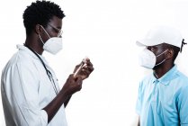 Médecin ethnique remplissant la seringue du flacon avec le vaccin se préparant à vacciner le patient afro-américain mâle en fond blanc dans une clinique pendant l'épidémie de coronavirus — Photo de stock