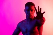 Serious young African American male athlete with naked torso looking at camera on pink background in neon studio — Stock Photo
