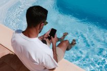 Vista posterior desde arriba de un hombre serio sentado junto a la piscina con las piernas en el agua y navegando en el teléfono móvil durante el trabajo remoto en verano - foto de stock