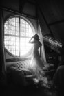 Черно-белый вид сзади на неузнаваемую женщину в платье, стоящую на диване напротив круглого окна в доме в солнечный день — стоковое фото
