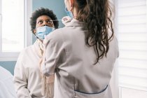 Специалистка-женщина в защитной форме, латексных перчатках и маске для лица делает тест на коронавирус носа у взрослой афроамериканской пациентки в клинике во время вспышки вируса — стоковое фото