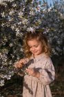 Liebenswert lächelndes kleines Kind im Kleid, das neben blühendem Baum mit Blumen im Frühlingspark steht und nach unten schaut — Stockfoto