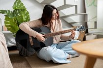 Giovane musicista donna suonare la chitarra acustica mentre seduto con le gambe incrociate sul pavimento a casa — Foto stock