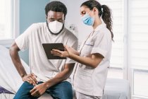Junge Ärztin in Arztuniform und Stethoskop mit Gesichtsmaske spricht mit afroamerikanischer Patientin während eines Termins in der Klinik und zeigt Ergebnis auf Tablet — Stockfoto