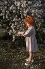 Adorable niño sonriente en vestido de pie cerca del árbol en flor con flores en el parque de primavera y mirando hacia abajo - foto de stock