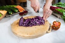 Обрезанная неузнаваемая женщина режет красную капусту ножом во время приготовления вегетарианской пищи за столом в лофт-хаусе — стоковое фото