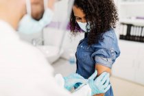 Cropped unrecognizable male medical specialist in protective uniform, latex gloves and face shield vaccinating African American female patient in clinic during coronavirus outbreak — Stock Photo