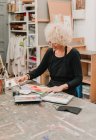 Зосереджена художниця сидить за столом і малює акварелями на папері під час роботи в творчій майстерні — стокове фото