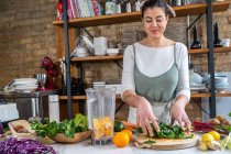 Female putting fresh chard leaves blender bowl with orange slices in house kitchen — Stock Photo