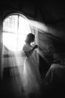 Чорно-білий вид невизначеної жінки в сукні, що стоїть на круглому вікні в будинку в сонячний день — стокове фото