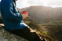 Вид сбоку на обрезанный неузнаваемый мужчина-турист, сидящий на холме с металлической чашкой горячего напитка и любующийся удивительными пейзажами горной местности в солнечный день — стоковое фото