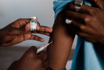 Médico negro en uniforme protector y guantes de látex vacunando a un paciente afroamericano irreconocible en la clínica durante el brote de coronavirus - foto de stock