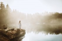 Voltar vista mulher de pé vestida com um vestido branco em uma rocha olhando para um lago em um dia nebuloso — Fotografia de Stock