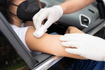 Cropped unrecognizable doctor in protective uniform, latex gloves and face shield disinfecting African American female patient arm with cotton and alcohol preparing to vaccinate inside the car on a drive through mobile clinic during coronavirus outbr — Stock Photo