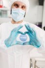 Positiver männlicher Arzt mit Latexhandschuhen und medizinischer Schutzmaske macht Herzensgeste auf Ich bin geimpft Botschaftsaufkleber auf weißer Uniform steht in moderner Arztpraxis und schaut in die Kamera — Stockfoto