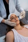 Cosmetólogo irreconocible recorte haciendo descamación facial ultrasónica para la mujer relajada acostada en la mesa médica en la clínica de belleza moderna - foto de stock
