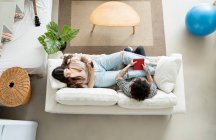 Hohe Winkel der jungen weiblichen SMS auf dem Handy, während auf der Couch mit tätowierten männlichen Geliebten zu Hause ausruhen — Stockfoto