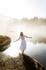 Back view woman standing dressed in a white dress on a rock looking at a lake on a foggy day — Stock Photo