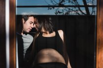 A través de la ventana de la mujer embarazada y el hombre contento tiernamente tocando las narices en el día soleado en casa - foto de stock