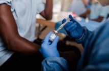 Schnitthände eines anonymen Arztes in Latex-Handschuhen füllen Spritze aus Flasche mit Impfstoff bereit, um unkenntlich männliche Afroamerikaner in Klinik während Coronavirus-Ausbruch zu impfen — Stockfoto