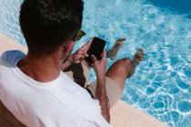 Сверху сзади вид серьезного мужчины, сидящего у бассейна с ногами в воде и просматривающего мобильный телефон во время удаленной работы летом — стоковое фото