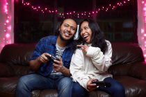 Возбужденная этническая пара в повседневной одежде с джойстиками, играющая в видеоигры вместе, сидя дома на кожаном диване — стоковое фото