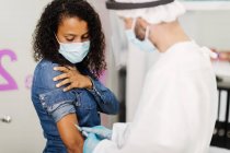 Побочный обзор неузнаваемого специалиста-мужчины в защитной форме, латексных перчатках и защитном щите лица, вакцинирующего афроамериканку во время вспышки коронавируса в клинике — стоковое фото
