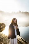 Donna in piedi vestita con un vestito bianco e giacca su di esso su una roccia guardando un lago in un giorno nebbioso — Foto stock