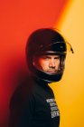 Вид збоку на впевненого брутального дорослого чоловіка в шоломі на чорному мотоциклі, який дивиться на камеру, стоячи на тлі барвистого червоного і жовтого фону — стокове фото