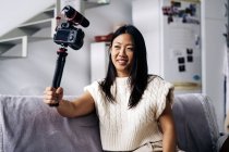 Улыбающаяся этническая женщина-блогер записывает видео на фотокамеру, сидя на диване в гостиной — стоковое фото