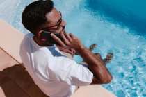 From above serious male freelancer sitting at poolside with legs in water and speaking on mobile phone during remote work in summer — Stock Photo