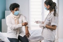 Especialista médica femenina en uniforme protector, guantes de látex y mascarilla facial vacunando a una paciente afroamericana madura en clínica durante el brote de coronavirus - foto de stock