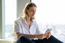Solitario unemotional giovane donna seduta in ufficio vuoto con grande finestra di navigazione sul telefono cellulare — Foto stock