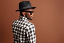 Giovane modello afroamericano maschile fresco in camicia a scacchi, cappello e occhiali da sole mentre in piedi su sfondo marrone — Foto stock