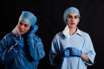 Young female coworkers in medical uniforms taking off face masks while standing on black background in clinic — Stock Photo