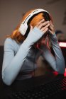 Side view of worried female gamer in headphones playing video game while sitting at table with keyboard — Stock Photo