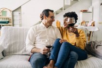 Zufriedenes multiethnisches Paar, das zu Hause mit Rotwein im Glas auf dem Sofa chillt, während es das Wochenende zu Hause genießt und sich gegenseitig anschaut — Stockfoto
