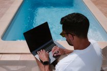 Freelancer masculino tumbado en una tumbona junto a la piscina y navegando por Internet en el portátil durante el teletrabajo en verano en un día soleado - foto de stock