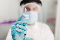Неопознанный медик в защитной маске и латексных перчатках с флаконом коронавирусной вакцины и шприцем, показываемым на камеру, стоя в больничной палате — стоковое фото