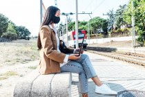 Бічний вид азіатки в масці сидить на лавці на залізничній станції і переглядає ноутбук під час очікування поїзда. — стокове фото