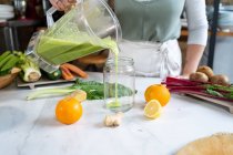 Crop fêmea anônima derramando saboroso smoothie de tigela de liquidificador em frasco à mesa com frutas cítricas em casa — Fotografia de Stock